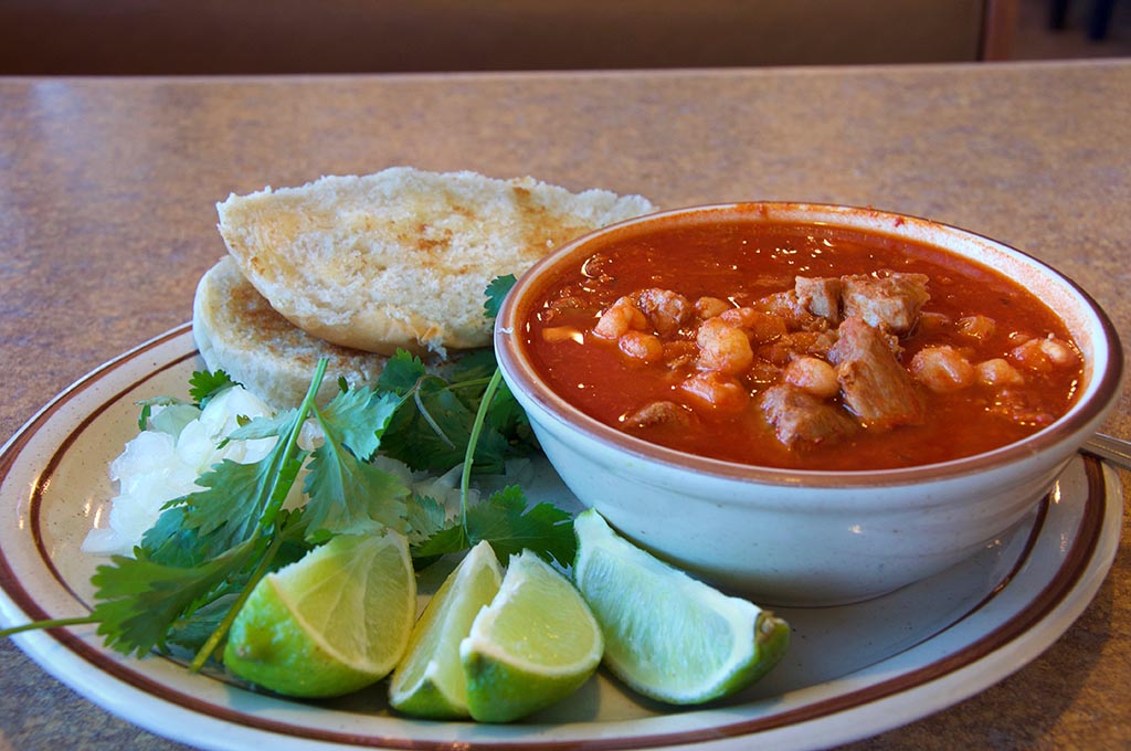 Bowl of red posole with limes, cilantro, onions and bread on the side.